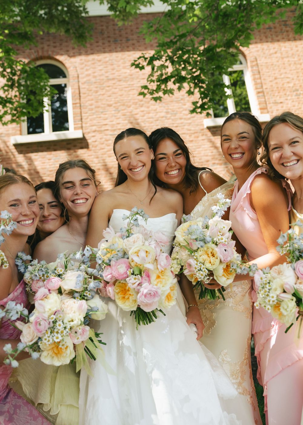 Smiling bride in white gown poses with bridesmaids in pink dresses holding floral bouquets outdoors near brick building.