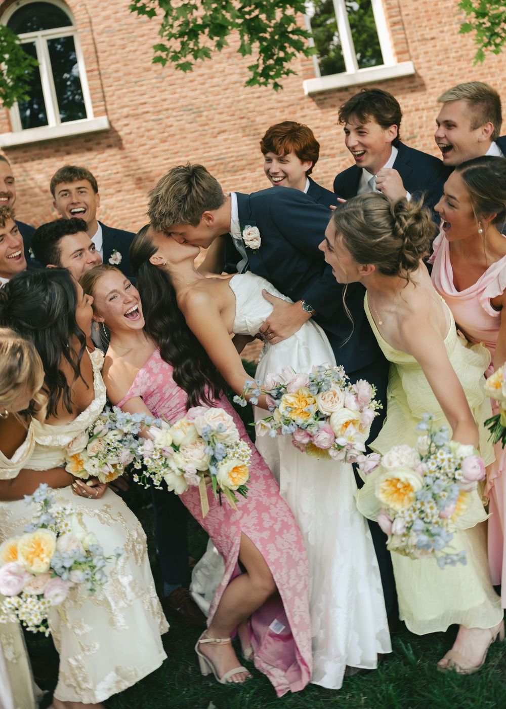 A joyful wedding party surrounds a couple sharing a kiss, with bridesmaids holding pastel bouquets outdoors.