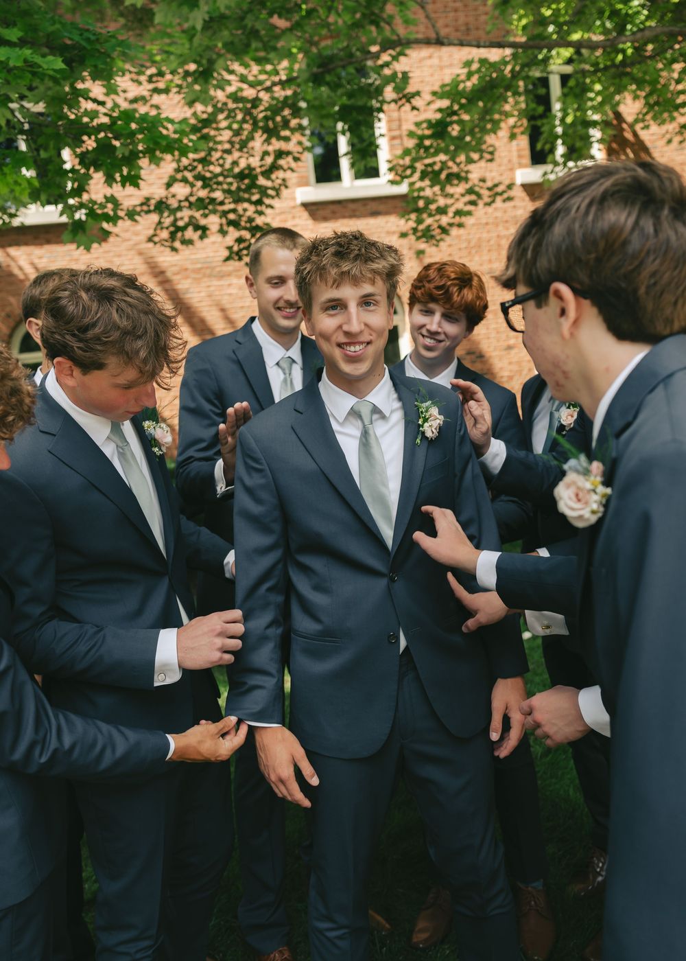 Groomsmen in gray suits with boutonnieres gather around smiling groom outdoors at wedding ceremony.
