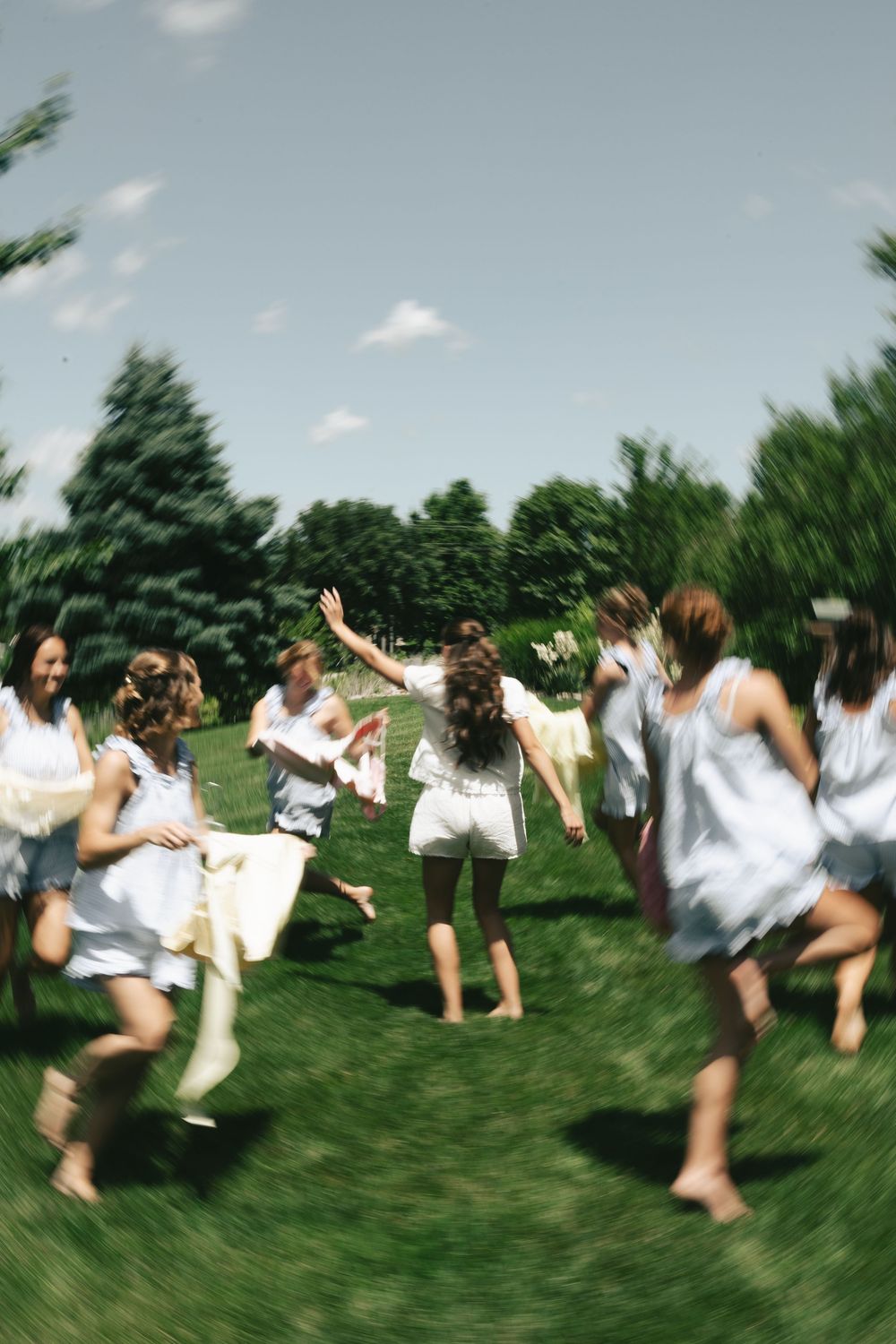 Women in white dresses running and celebrating outdoors on green grass surrounded by trees on a sunny day.