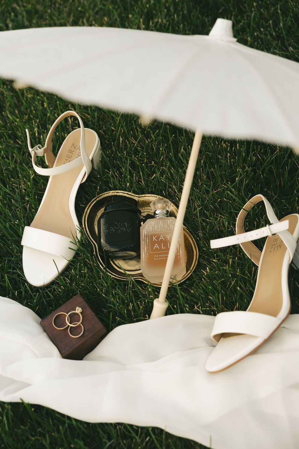 Bridal flat lay with white heels, gold perfume, rings, and parasol on green grass.