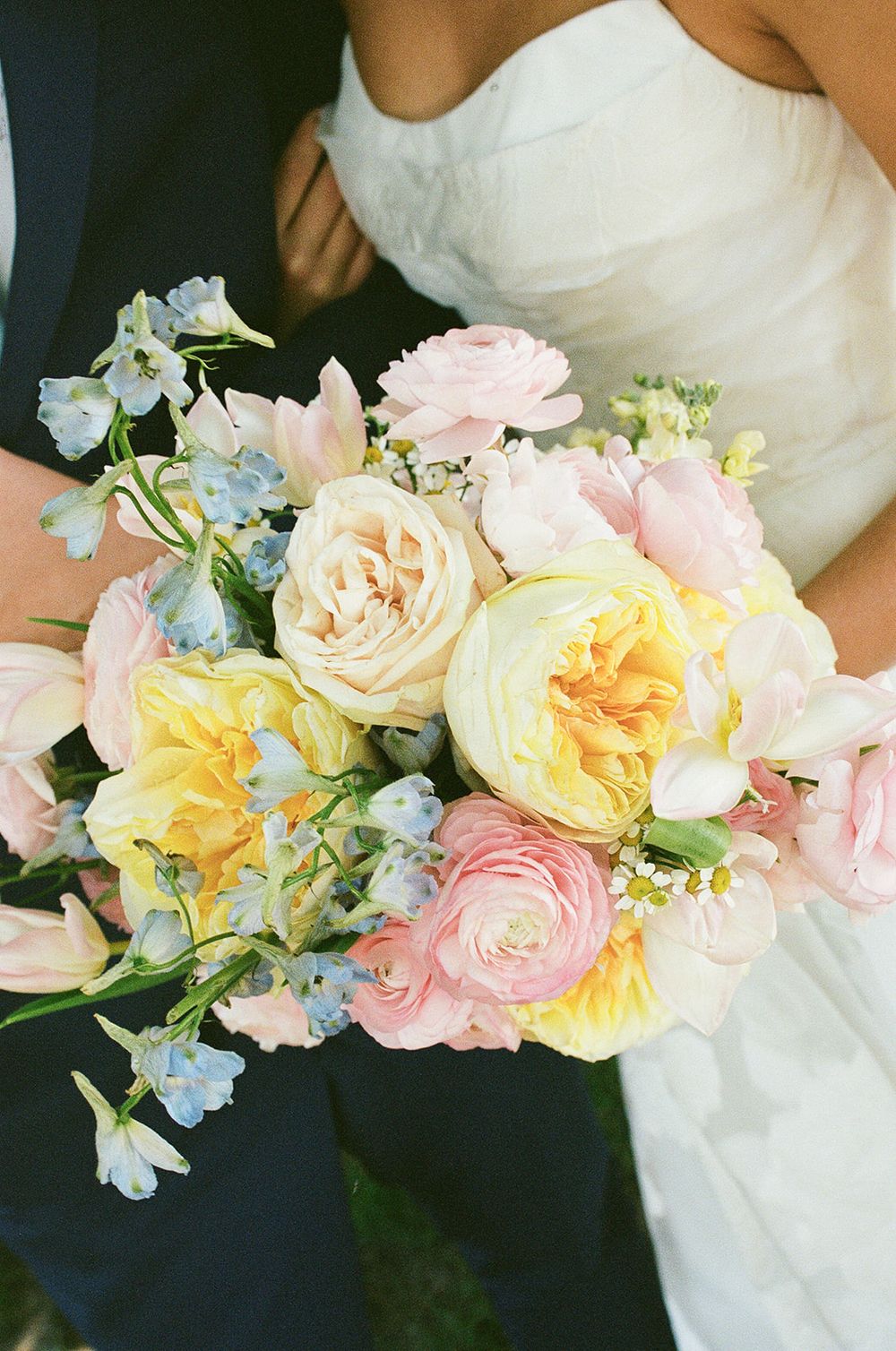 Bridal bouquet featuring yellow garden roses, pink ranunculus, and blue delphinium held by a couple in wedding attire.