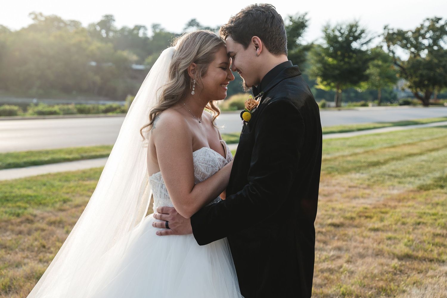 A couple shares an intimate moment in formal wedding attire on a grassy field during sunset.