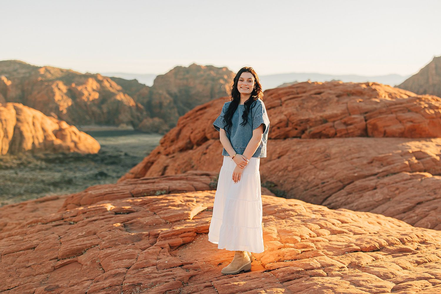 Woman in denim top and white skirt standing on red sandstone rocks with scenic canyon landscape.