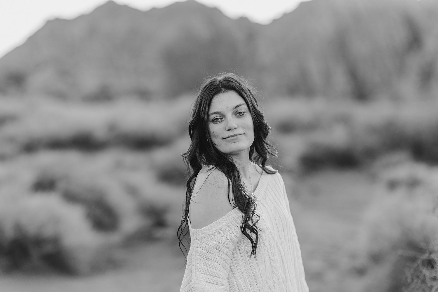 Black and white portrait of a young woman with long wavy hair in a white dress, posing outdoors with mountains behind.