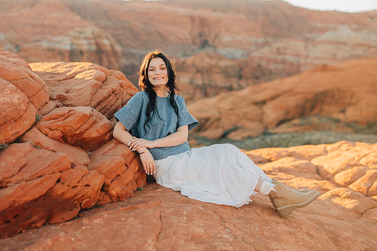 Smiling young woman in teal top and white skirt sits on red sandstone rocks in desert canyon.