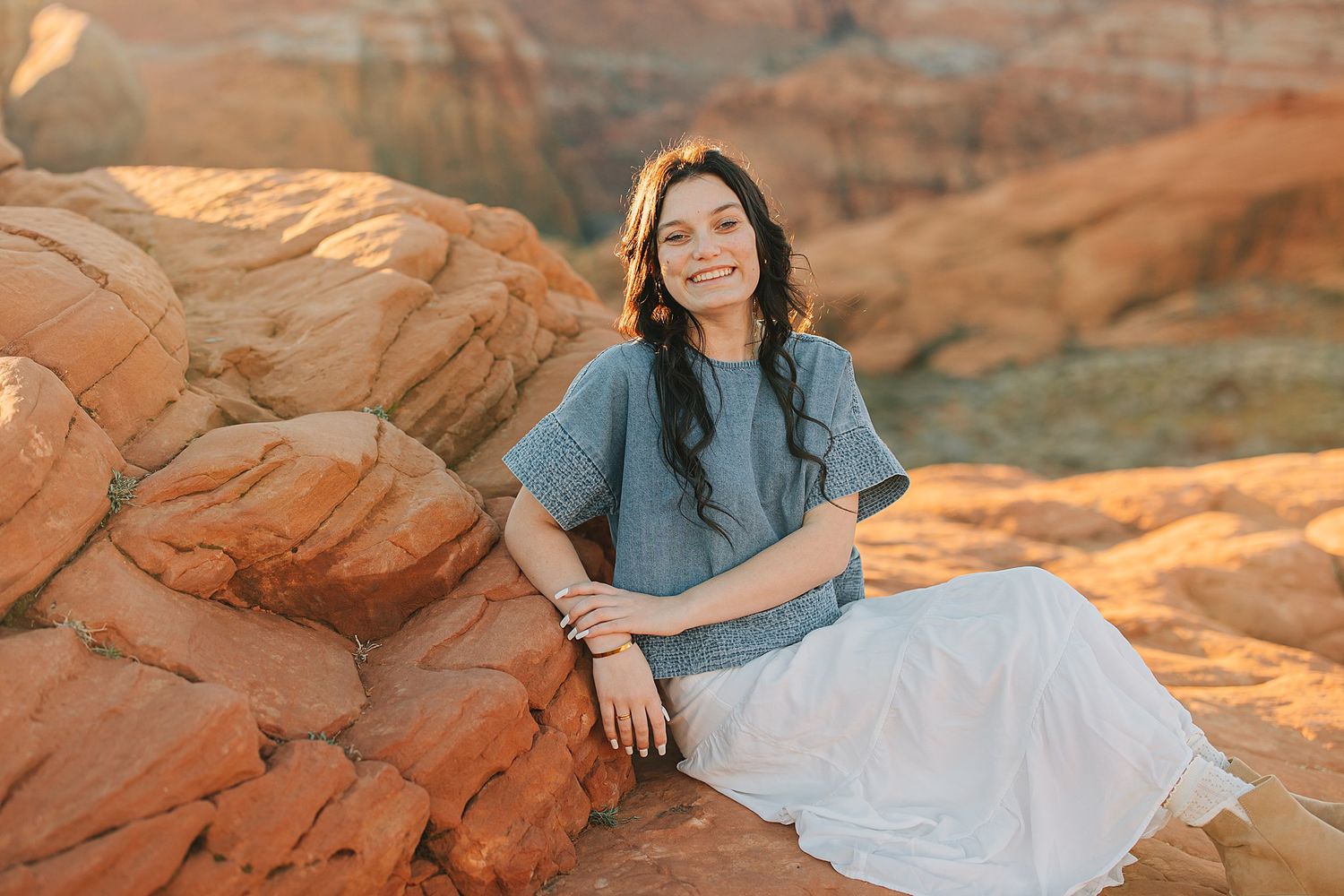 Woman in blue top and white skirt smiling on red rock formations in a desert landscape.