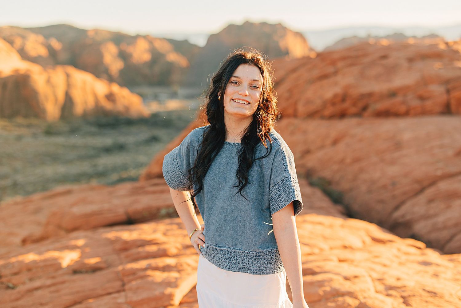 Young woman with dark hair smiling in desert landscape with red rock formations at golden hour.