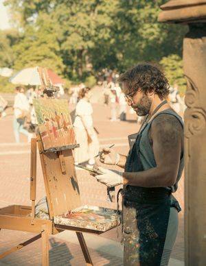 Male artist painting on an easel outdoors in a busy park plaza on a warm day.