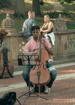 Young man playing cello outdoors near Bethesda Terrace in Central Park on a sunny day.