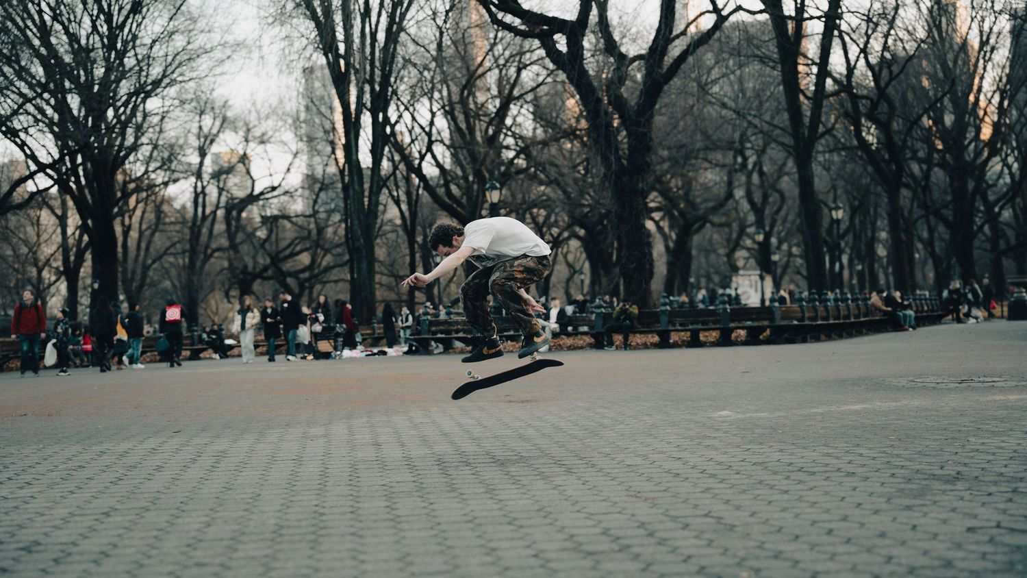 Skateboarder performing a jump trick in a busy Central Park plaza surrounded by spectators.