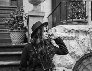 Black and white photo of a stylish woman in a wide-brim hat and coat posing outdoors near stone steps.