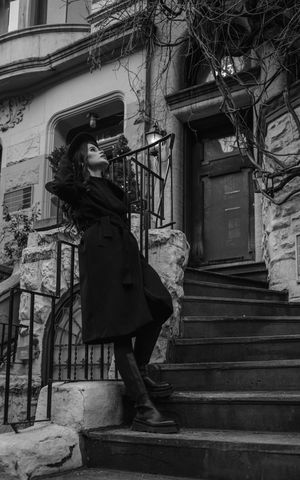Black and white photo of a woman in a black coat and hat posing on stone steps outside a vintage building.