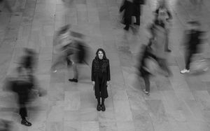 Black and white photo of woman standing still amid blurred moving crowd indoors.