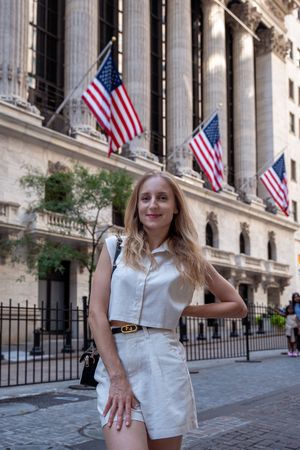 Blonde woman in white outfit posing in front of the New York Stock Exchange with American flags.
