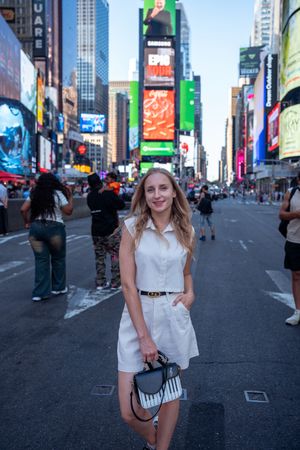 Smiling blonde woman in white dress posing in Times Square with bright billboards behind her.