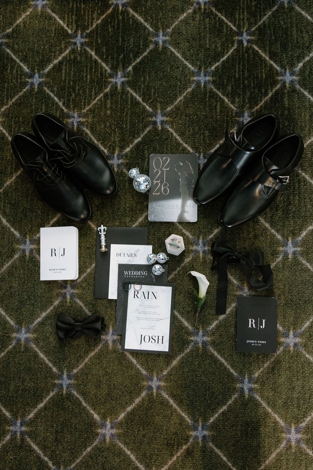 Wedding accessories including black dress shoes, invitations and decor arranged on a geometric patterned green carpet.