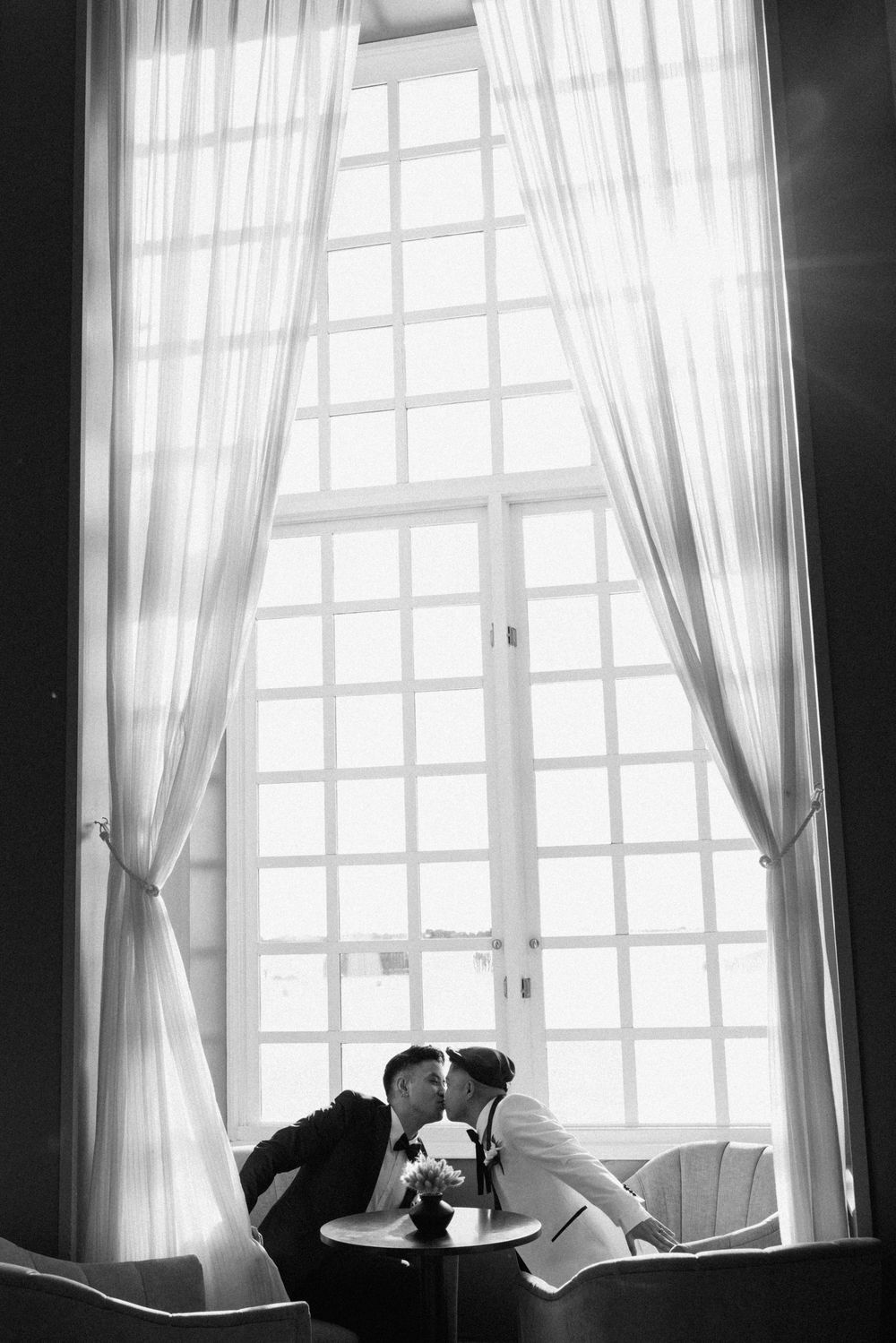 Silhouetted figures sit by bright windows with sheer curtains in a black and white interior scene.