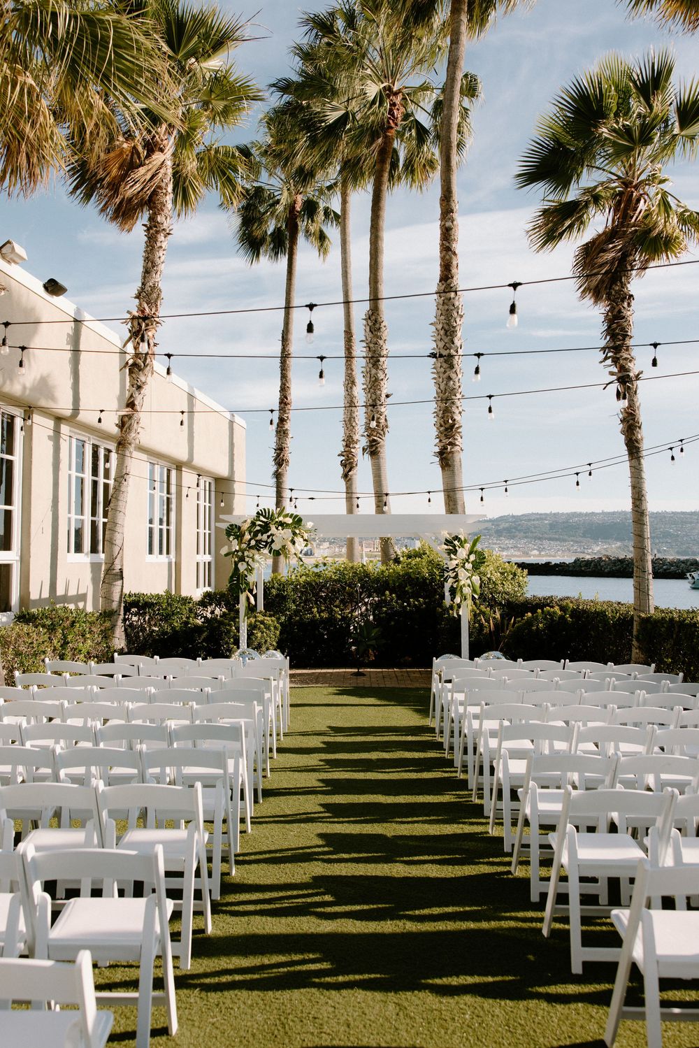 Outdoor wedding ceremony setup with white chairs and palm trees overlooking the water, with string lights hung overhead.