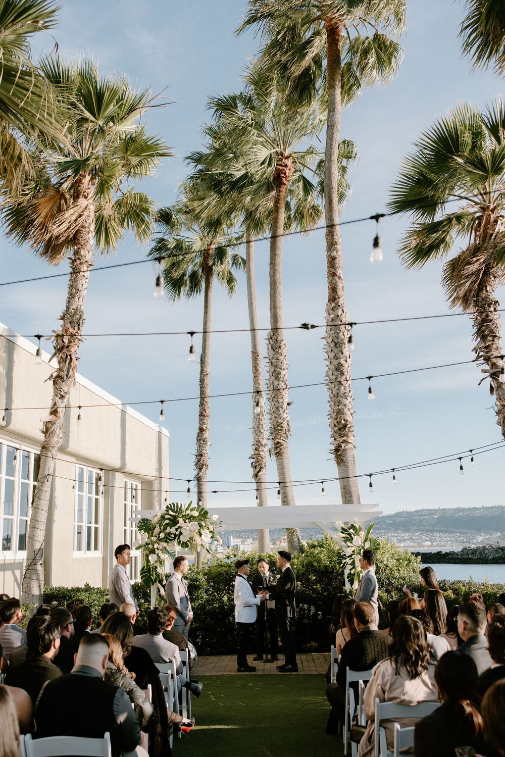Outdoor wedding ceremony setup with tall palm trees and string lights against a waterfront backdrop.
