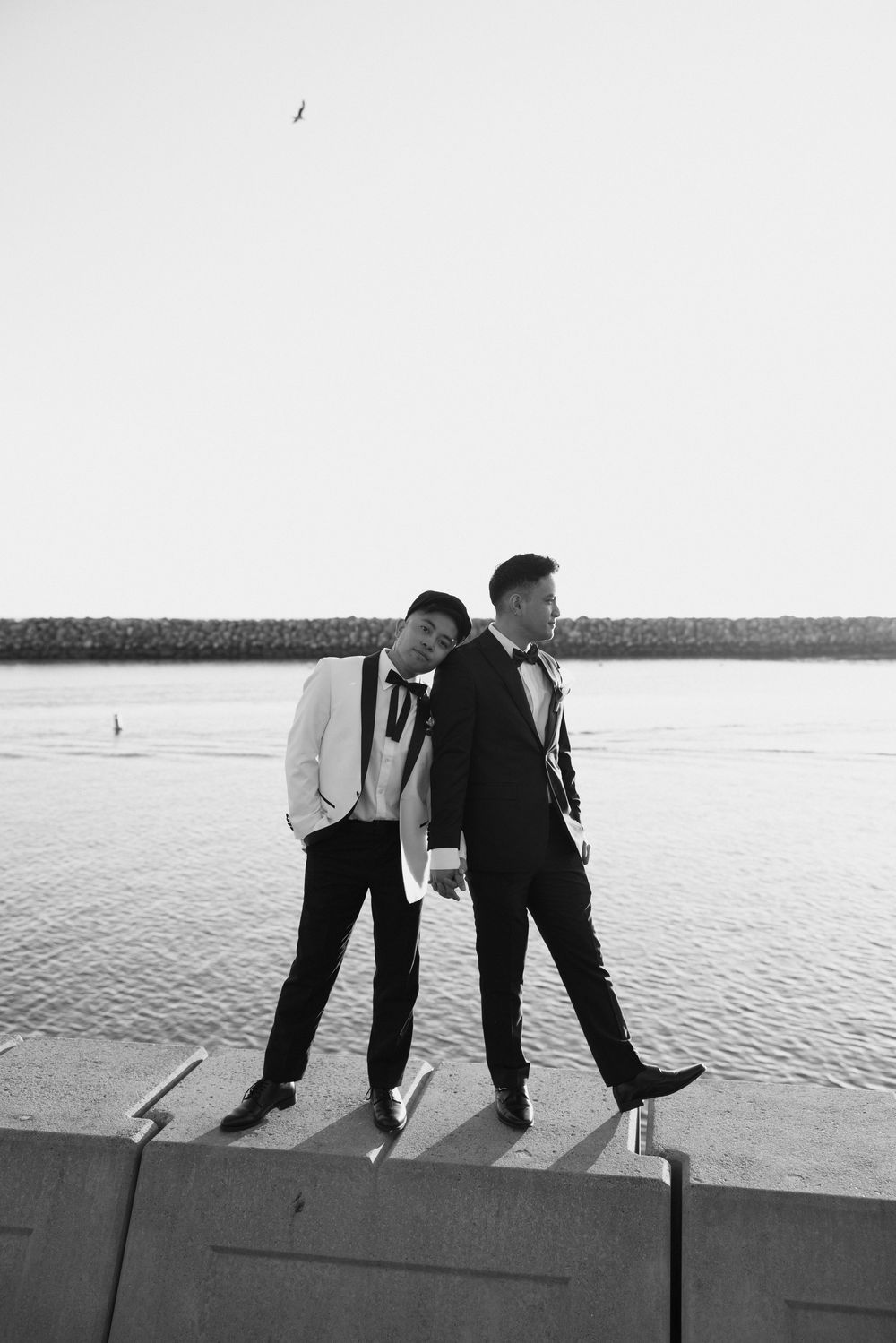 Two figures in formal attire playfully posing on a waterfront ledge at sunset.