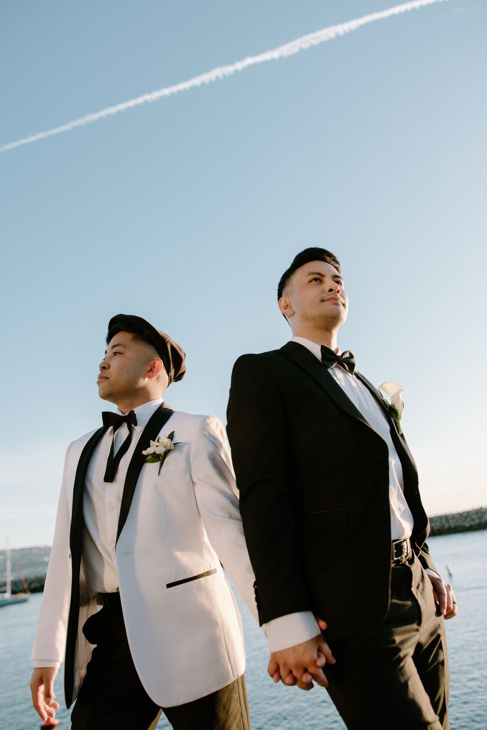 Two grooms in contrasting black and white tuxedos pose together against a bright blue sky with a jet stream.