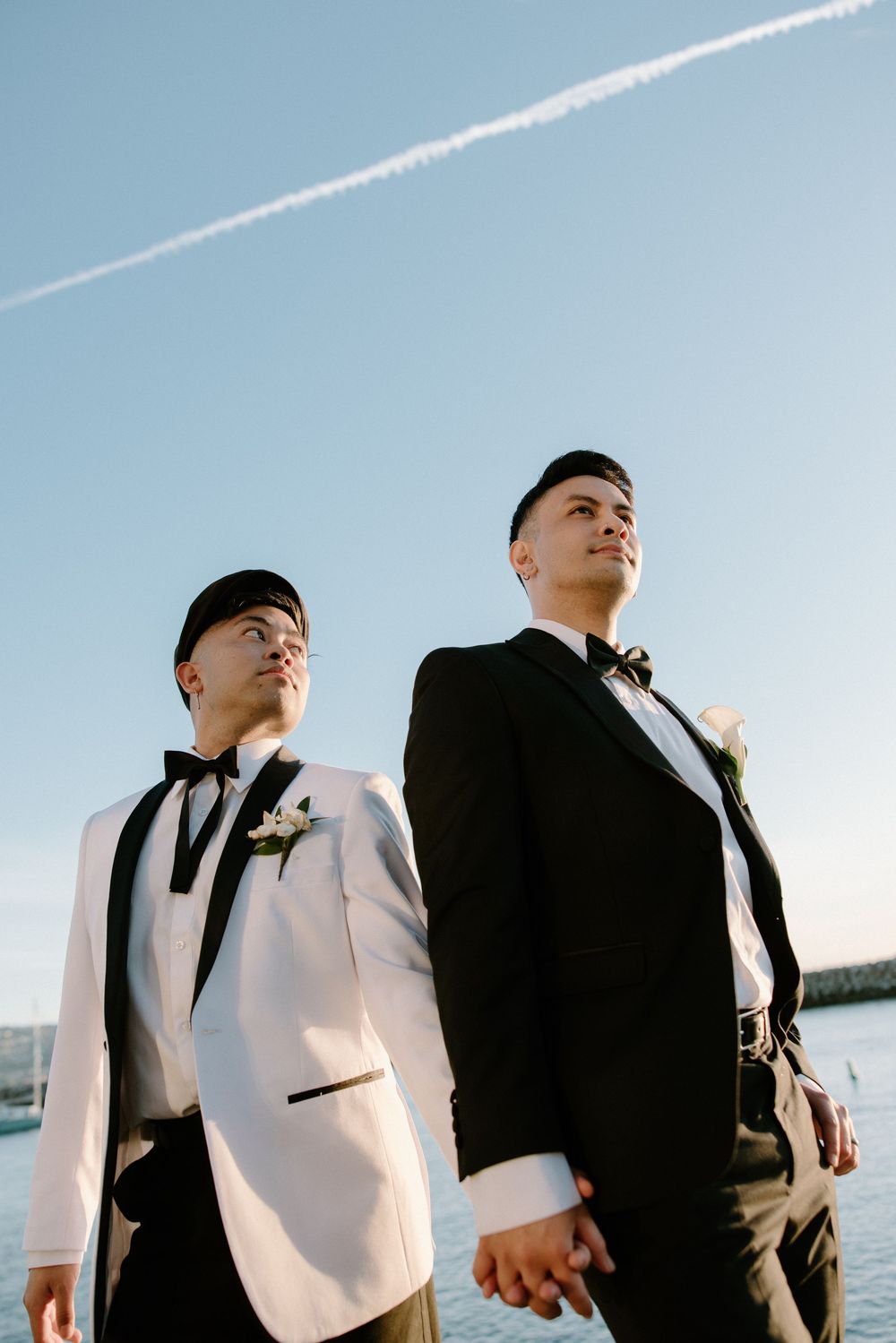 A couple in formal attire stands together against a waterfront backdrop on their wedding day.