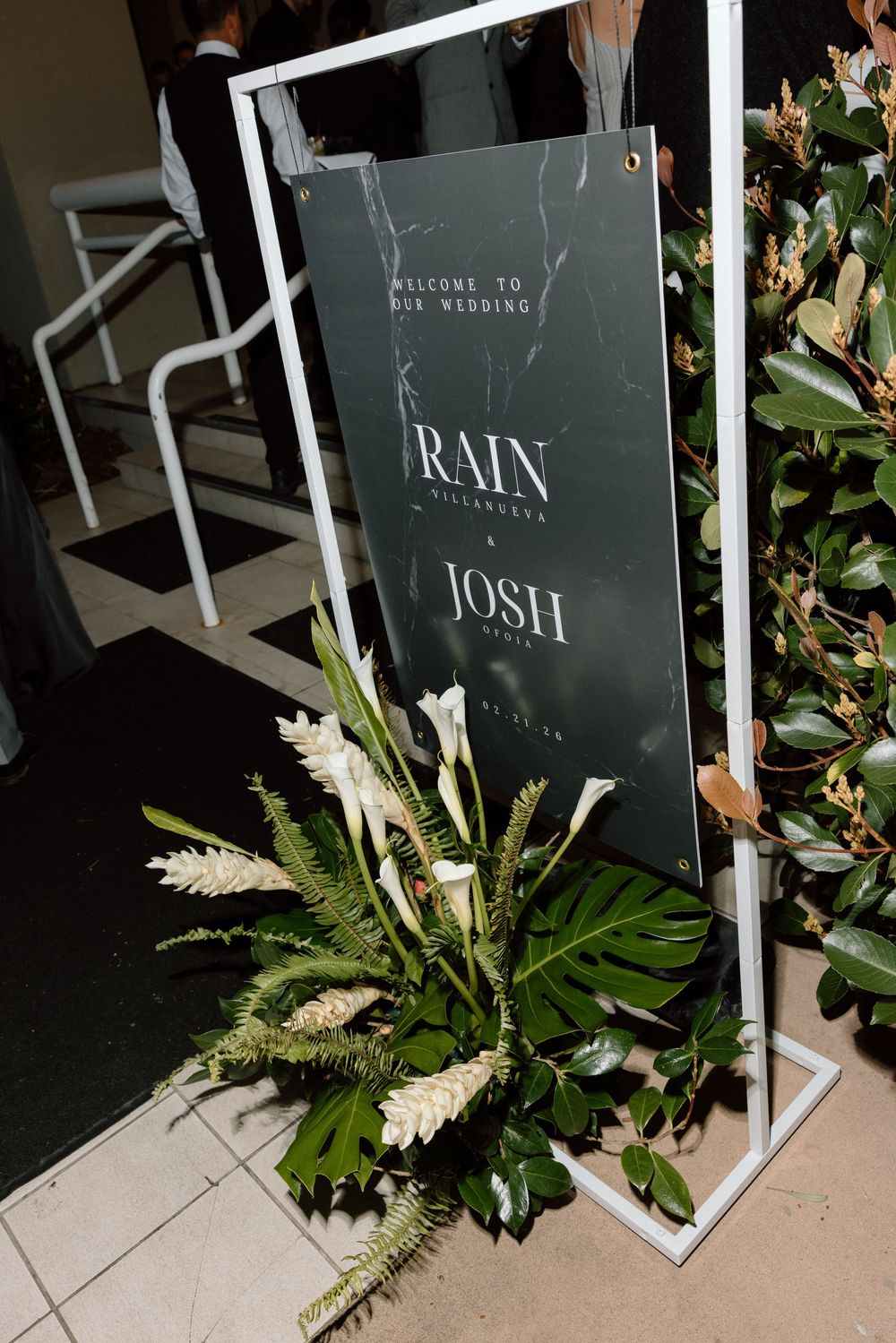 Modern wedding welcome sign with 'Rain & Josh' text surrounded by elegant white floral arrangements.