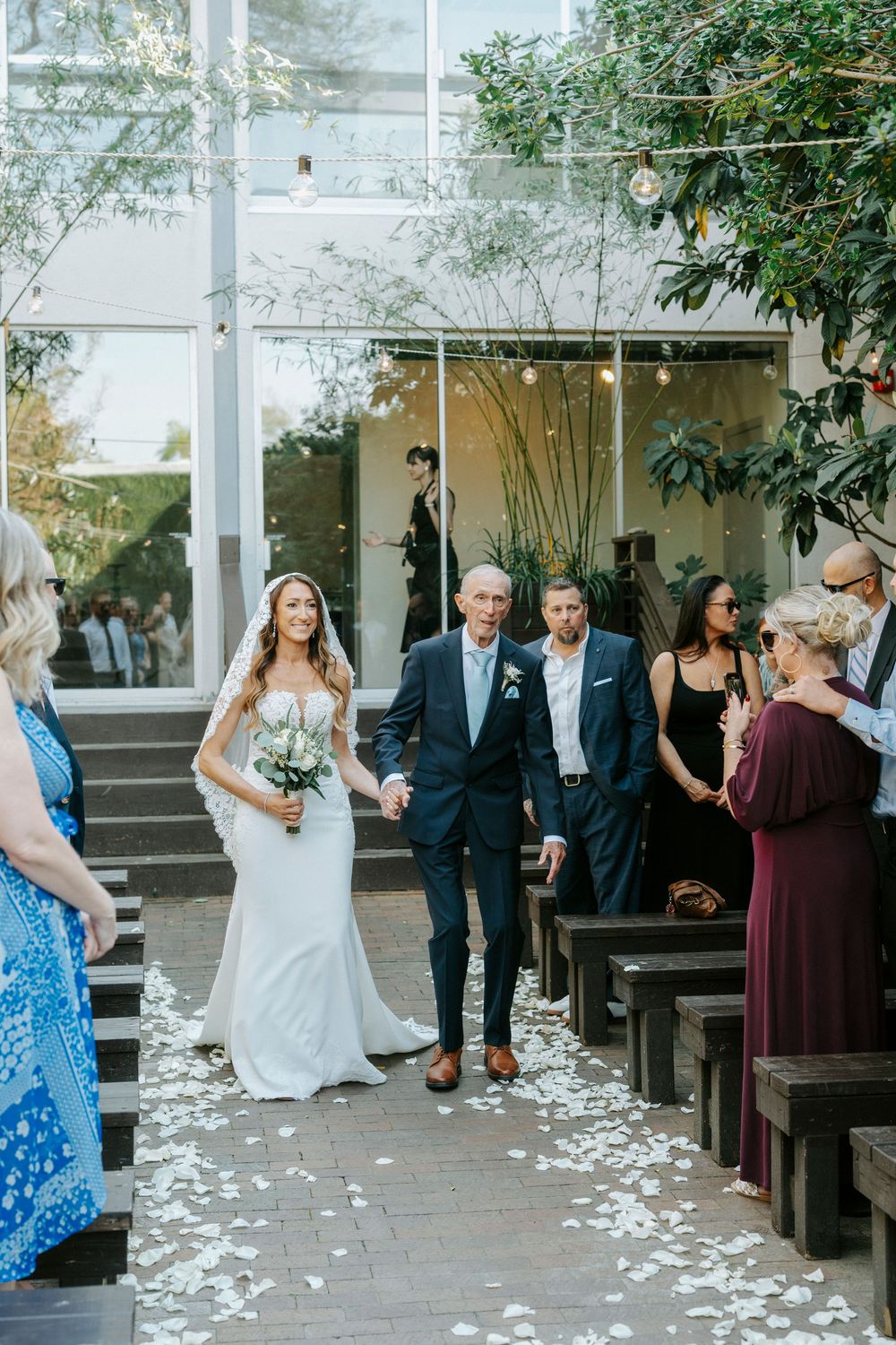 Wedding ceremony setup with white petals scattered along a brick pathway surrounded by greenery and columns.