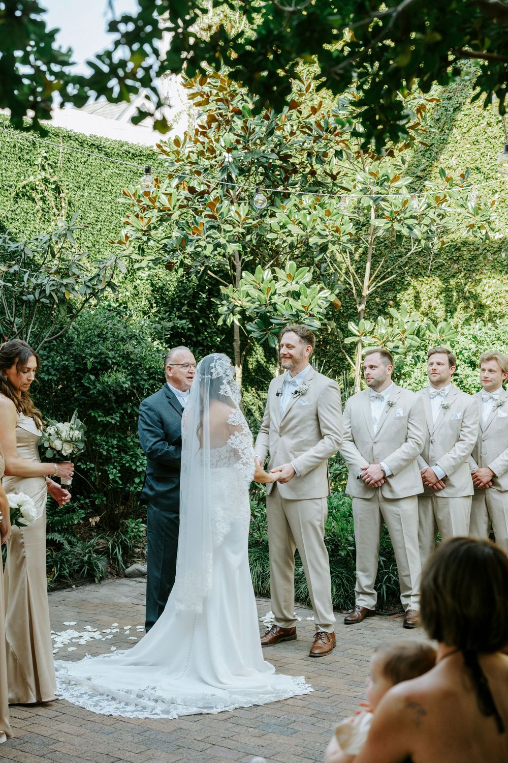 Wedding party in light beige suits and white dress gather outdoors on brick patio during garden ceremony.