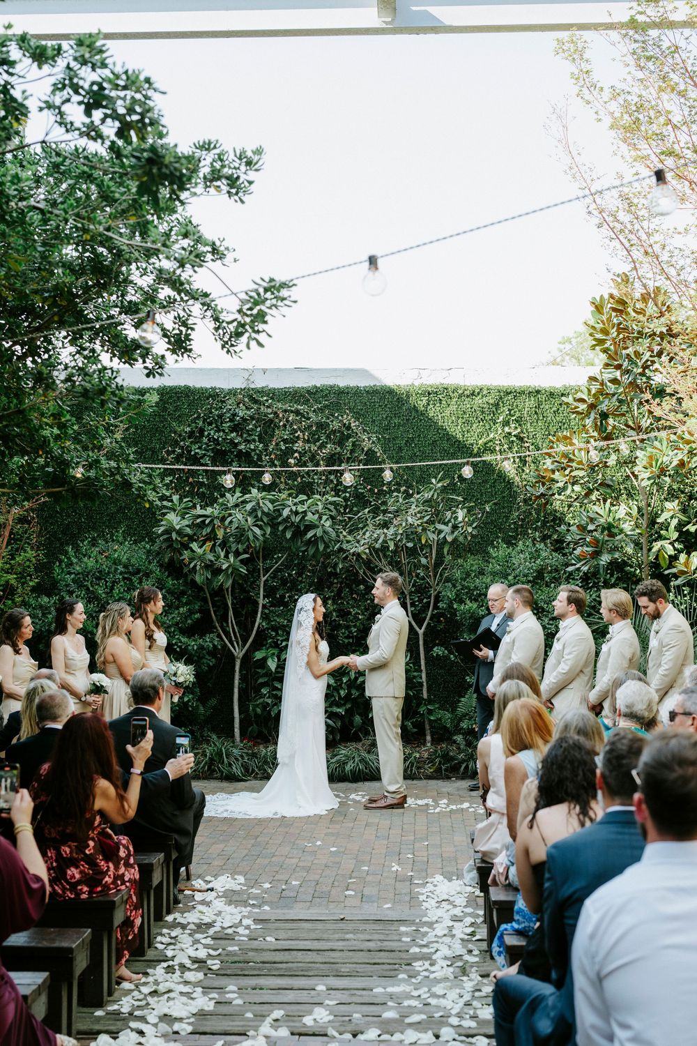 Outdoor wedding ceremony on a stone patio surrounded by trees with white flower petals scattered along the aisle.