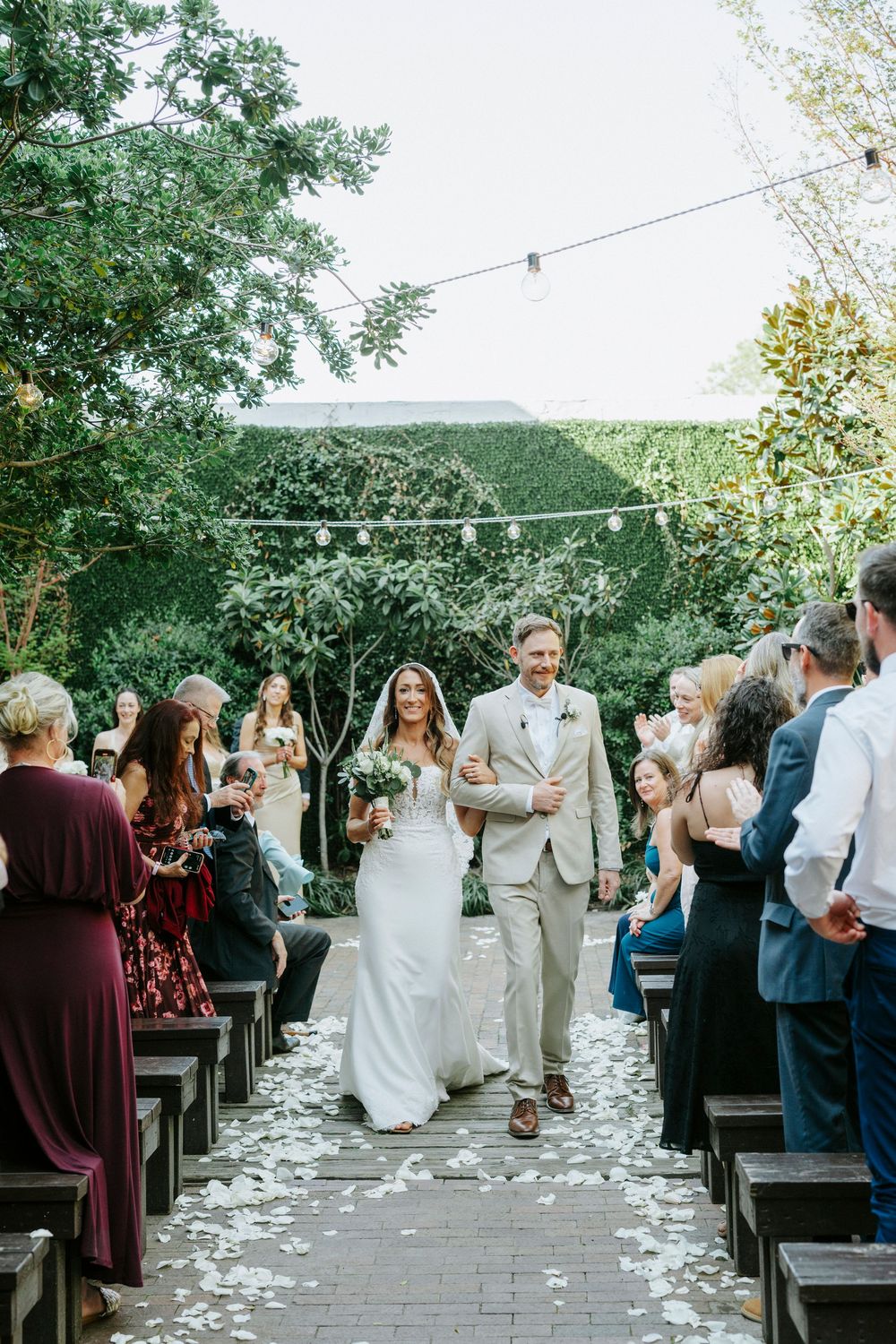 Wedding ceremony processional with attendees in burgundy dresses walking down a petal-lined aisle outdoors.