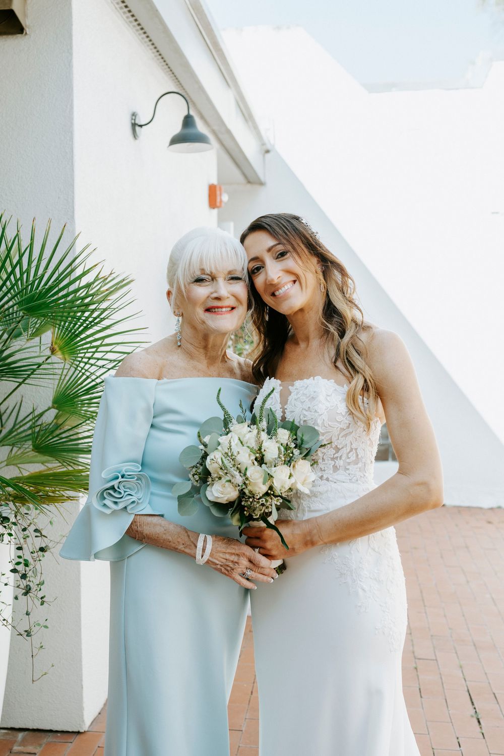 Two people in formal attire share an emotional moment while holding a white floral bouquet outdoors.