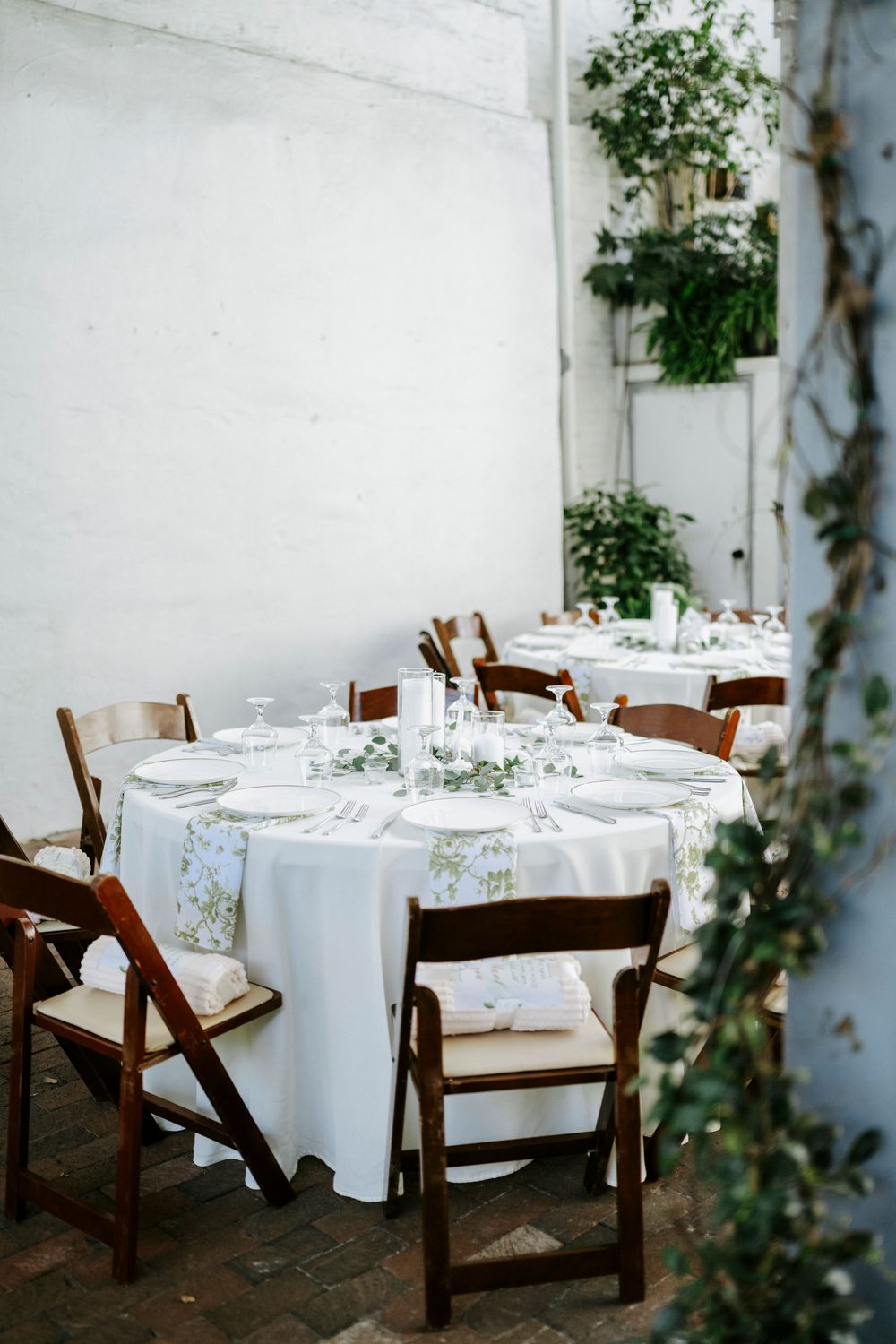 Elegant wedding reception table with white linens, wooden folding chairs and delicate floral arrangements against a white wall.