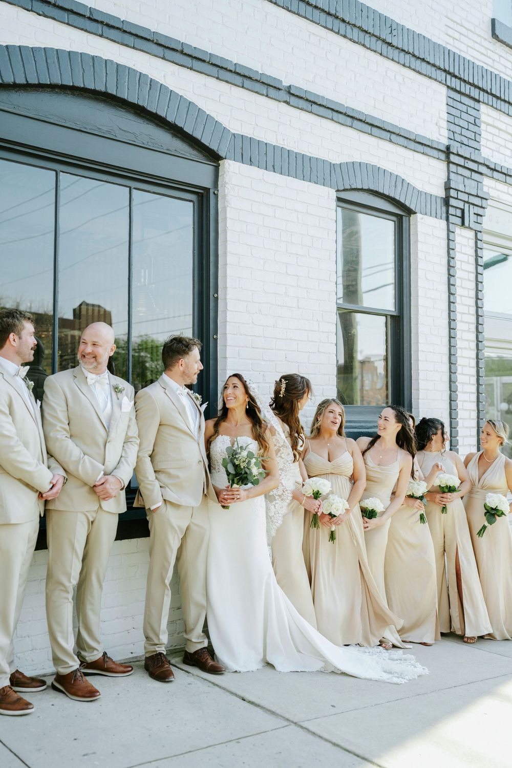 Wedding party in cream colored attire poses against modern white building with large windows.