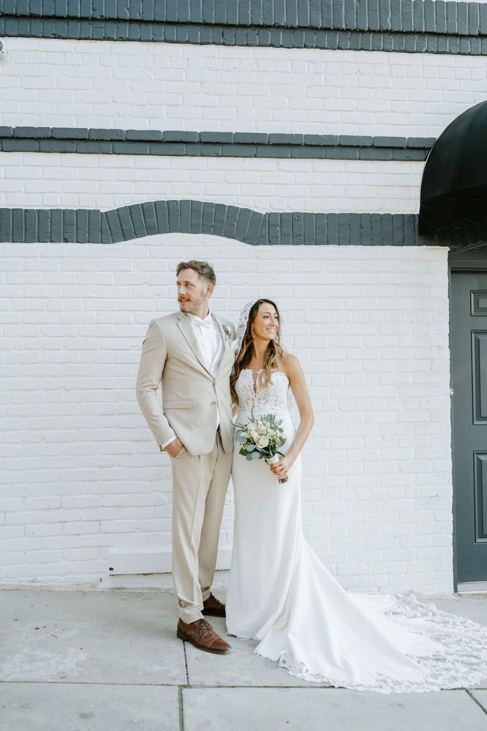 Elegant wedding portraits against a white brick wall featuring a beige suit and flowing white gown.
