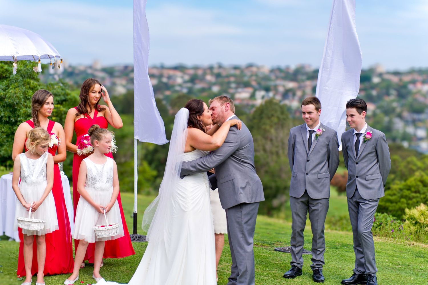 Wedding party on a grassy hilltop overlooking a scenic view, where the couple is kissing each other holding tight.