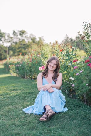 A person in a light blue dress sits among blooming flowers in a garden setting.