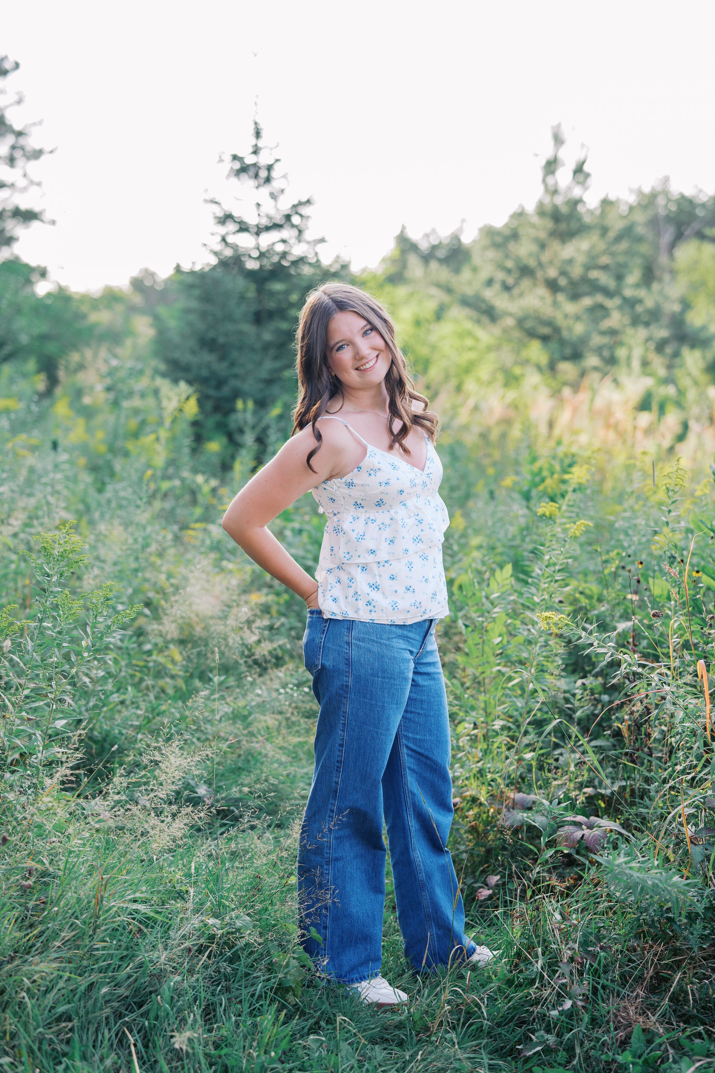 Full-length outdoor portrait in a meadow with evergreen trees in background.