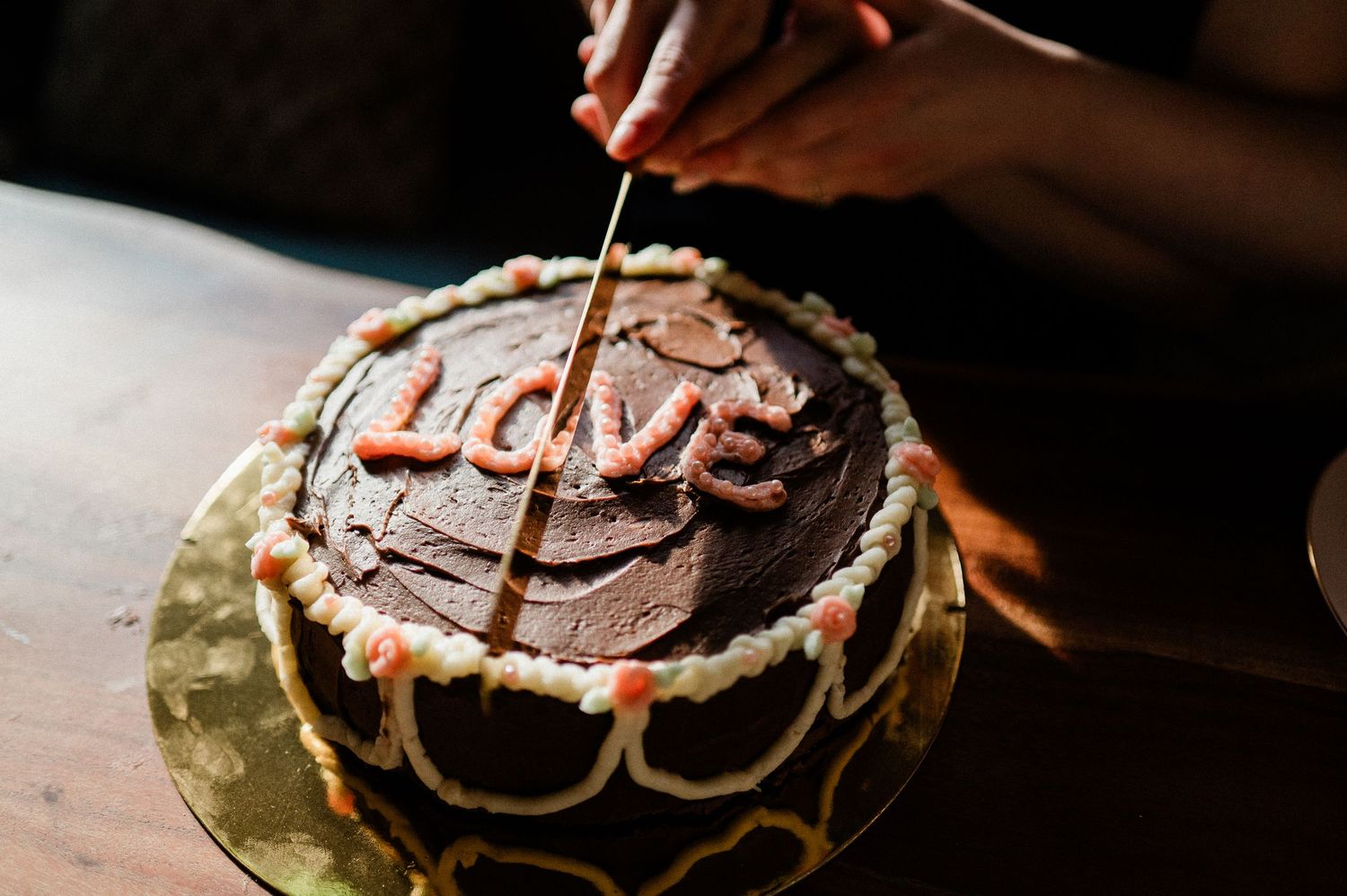A chocolate cake decorated with pink frosting and the word 'love' is being sliced on a wooden surface.