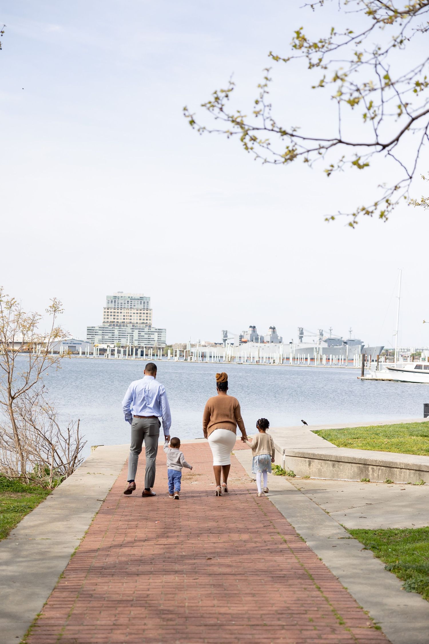 A family of four walks hand-in-hand along a brick waterfront path toward a scenic harbor on a clear day.