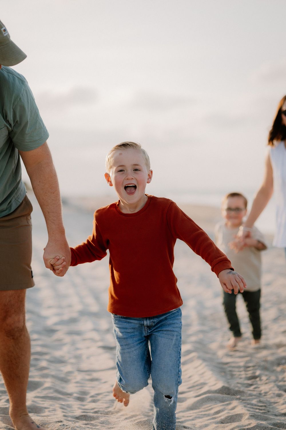 Family walking together on sandy beach at sunset with joyful expressions and casual attire.