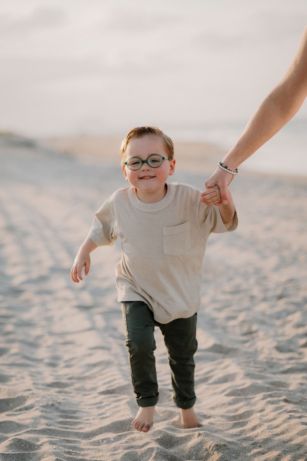 A small child wearing glasses and beige shirt holds an adult's hand while walking barefoot on sandy beach at sunset.