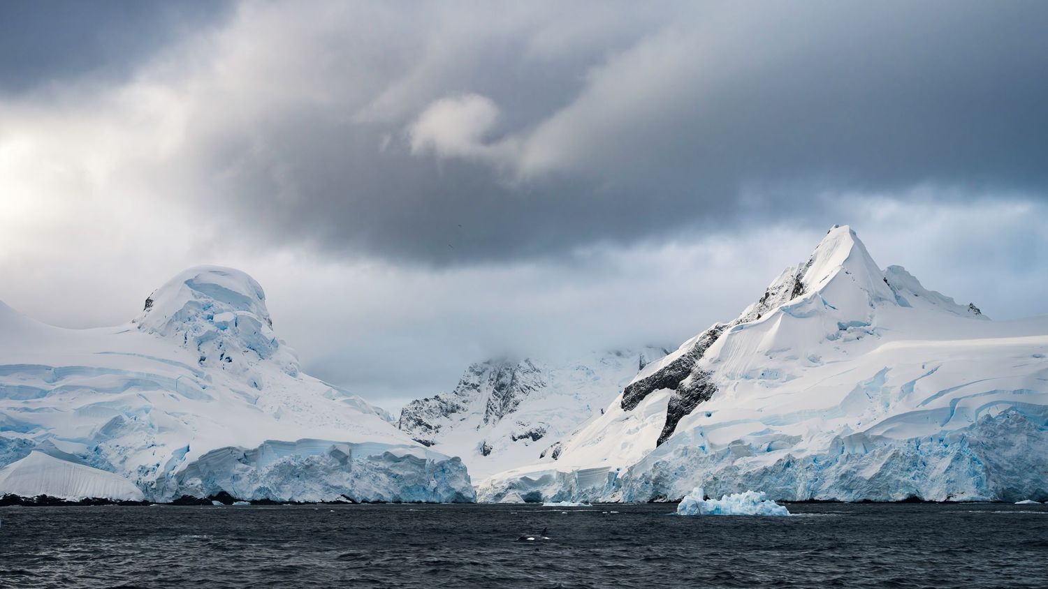 A panoramic view of snow-covered mountain peaks and glaciers under dramatic cloudy skies in Antarctica.