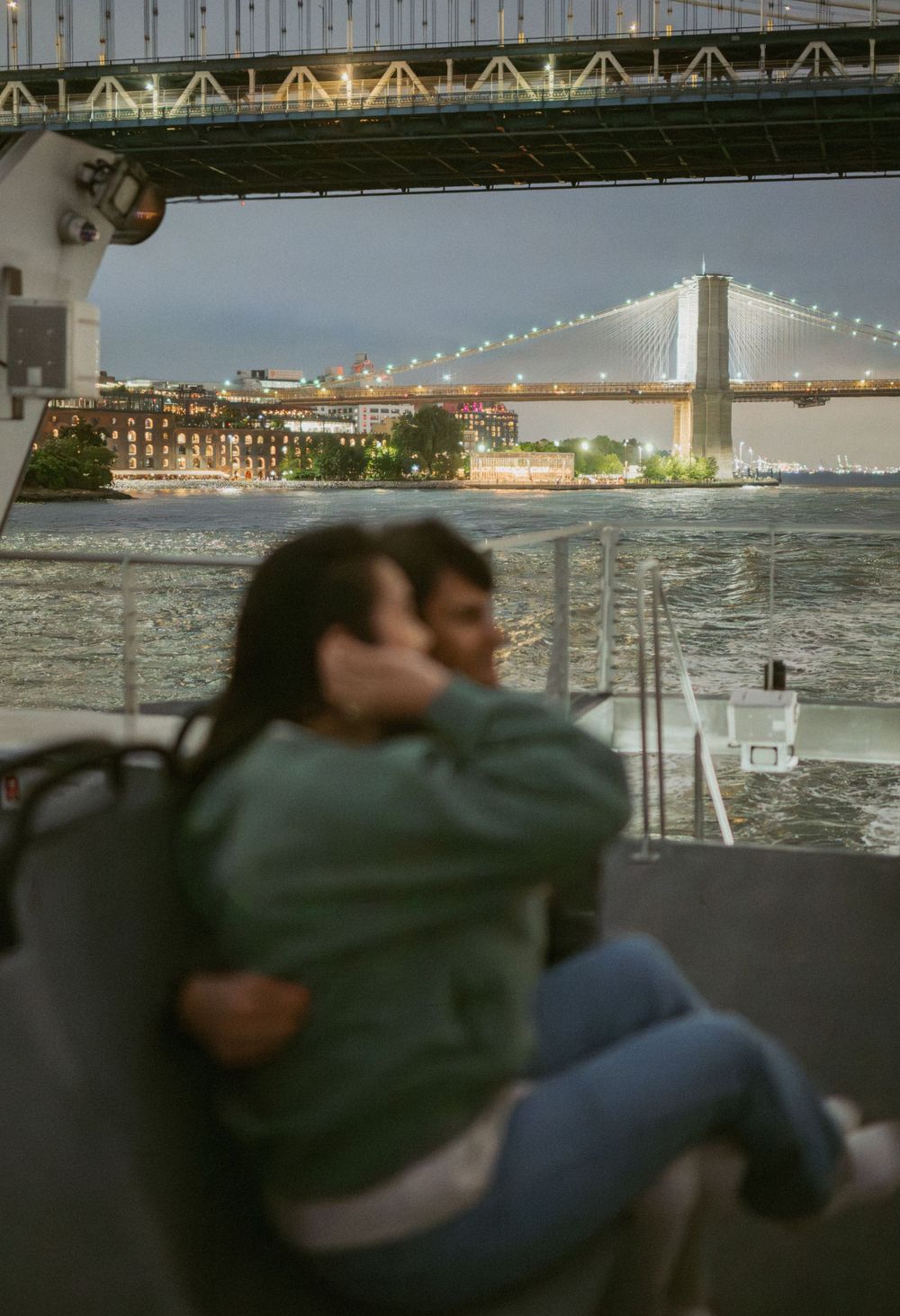 Couple embraces while looking at illuminated city skyline and bridge across water at dusk.