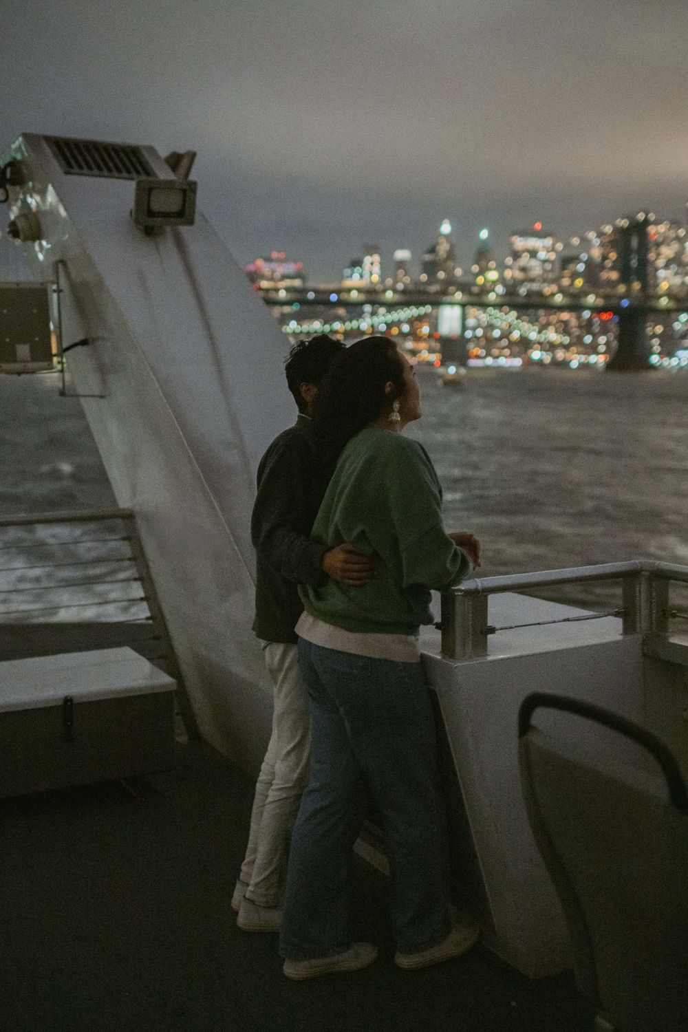 Two people lean against a railing overlooking a city skyline at night with twinkling lights in the distance.