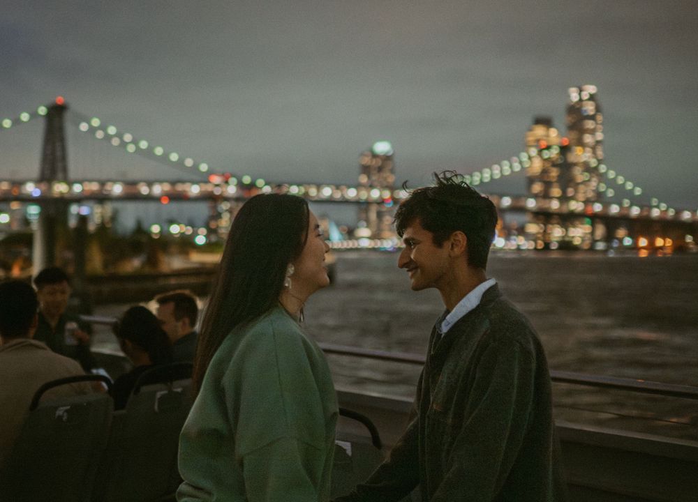 People gathered on waterfront viewing deck with twinkling city lights in background at night.