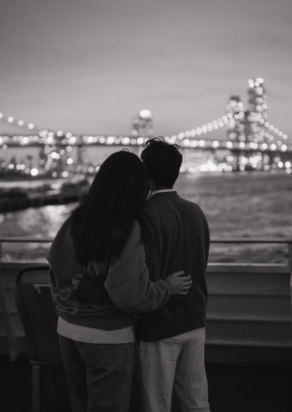 A couple embraces while looking at the illuminated city bridge lights reflecting over dark waters at night.