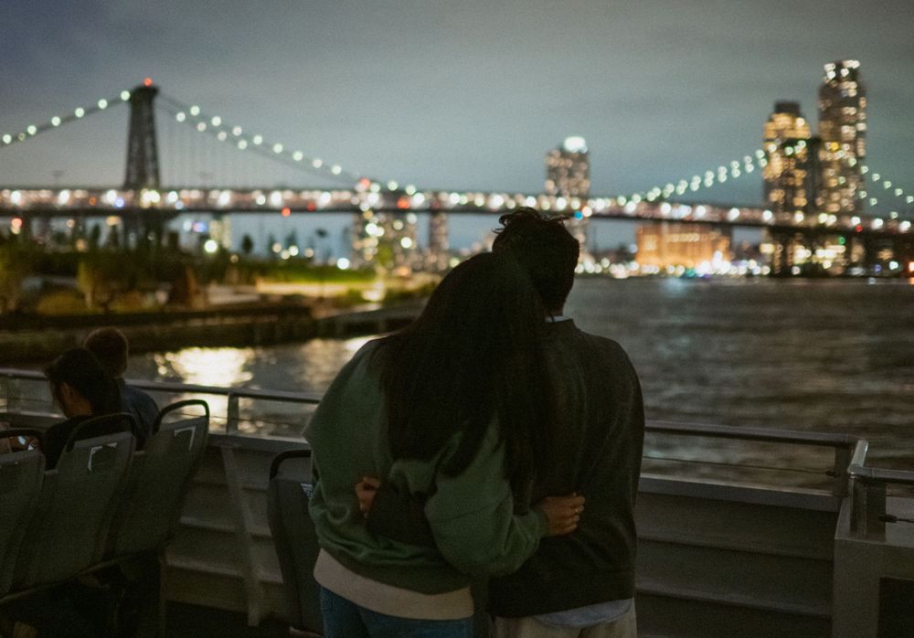 String lights illuminate a waterfront patio area with city skyline views at night.