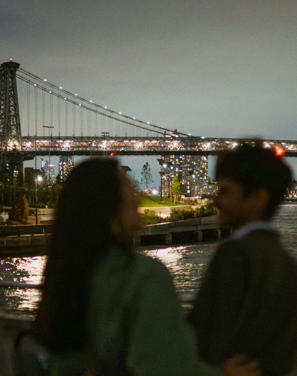 Silhouettes against the illuminated Williamsburg Bridge at night in New York City with glowing lights reflecting on water.